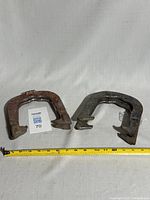 Two vintage forged iron horseshoes side by side with measuring tape in front showing size and scale.