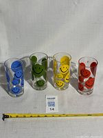 Front view of four drinking glasses arranged side by side with colorful happy face designs in blue, green, yellow, and red. A tape measure shows approximate height around 5.5 inches.