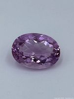 Close-up photo of an oval faceted purple amethyst gemstone against a light background.