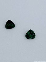 Two dark green demantoid garnet gemstones on white background, one trilliant cut and one heart shaped.