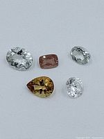 Five loose faceted gemstones of various shapes and colors laid out on a flat surface against a light grey background.