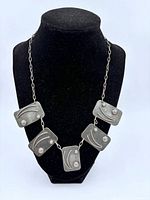 Front view of modernist metal link necklace on black display stand showing five rectangular dark silver tone metal pieces with abstract curved and dot designs connected by chain links.