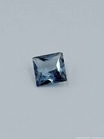 Close-up photo of a loose, square princess cut sapphire with visible facets and a bluish-gray hue on a white background.