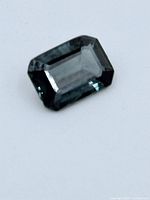 Close-up image of the sapphire gemstone on a white background showing its rectangular emerald cut and blue-green color.