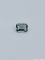 Front view of the rectangular emerald-cut bluish green Spinel gemstone on a light gray background.
