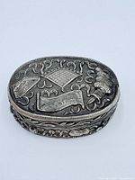 Top view of oval sterling silver pill case showing raised decorative motifs of a torch, banner, checkerboard table, and tassels.