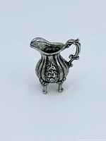 Front view of the sterling silver miniature pitcher showing floral pattern and three small feet.
