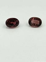 Two oval cut reddish-pink tourmaline gemstones shown side by side on white background.