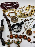 Photo showing an assortment of jewelry including necklaces, watches, bracelets, pendants, and pins placed on a white background.