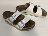 Side view of white Birkenstock sandals showing leather straps, metal buckles, cork footbed, and sole with visible wear and dirt.