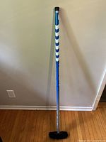 Full length image of the curling broom showing the handle, shaft with blue and white chevron grip, and black broom head on wooden floor against a wall.