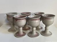Set of six pink and beige speckled stoneware chalices standing in a group on a white surface under natural light.