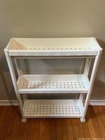Straight view of the white IKEA Vesken rolling shelf cart showing three perforated shelves on wooden floor and beige wall background.