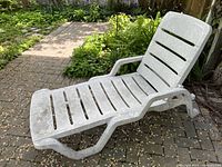 White plastic patio lounge chair positioned on a brick pathway with greenery in the background, showing the chair in a reclining position. Surface dirt and discoloration are visible.