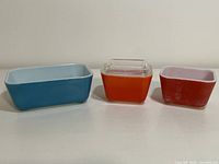 Front view of three rectangular vintage Pyrex Fridgie dishes in red, blue, and pink with clear glass lid on white surface