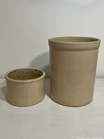 Photo of small and large beige stoneware crocks side by side on a white surface against white background.