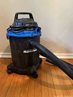 Full view of Mastervac wet/dry vacuum on caster wheels with attached hose on a wooden floor.