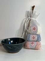 Photo showing pottery yarn bowl in dark blue with carved leaf design and spiral yarn guide holes, alongside packaged balls of cotton yarn in pastel colors and knitting needles.