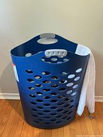Full view of the dark blue Rubbermaid laundry basket with multiple round ventilation holes and built-in handles. White Woolite mesh laundry bag partially visible inside or alongside the basket.