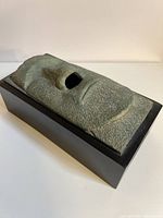 Front angled view showing textured Easter Island Moai head with tissue dispensing hole through nose area, mounted on black rectangular base.