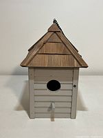 Front view of birdhouse showing circular entrance hole, small perch, white wooden siding and natural wood shingled roof with black chain on top.
