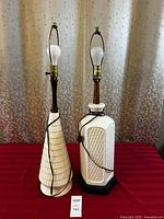 Pair of two vintage white/cream ceramic table lamps with dark wood necks and brass fittings, on red fabric against floral curtain backdrop.