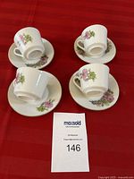 Four white porcelain espresso cups and matching saucers with purple floral patterns and gold trims, arranged on a red cloth.