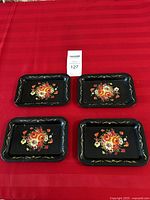 Four small black metal trays with floral paintings arranged on red fabric
