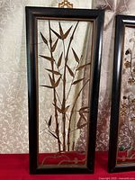 Front view of a black framed wood and bronze metal bamboo plant art piece showing detailed metal leaves and stalks within an open back frame.
