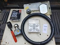 Photo showing a black leather-textured steering wheel cover, chrome McGard wheel locks with key, Fram oil filter wrench, and other automotive items on a white board.