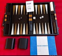 Open backgammon board inside folding case on red surface showing playing chips arranged on right, dice, doubling cube, and instruction booklet beneath.