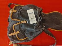 Black canvas knapsack with tan leather trim and silver buckles shown front and angled view on red surface.