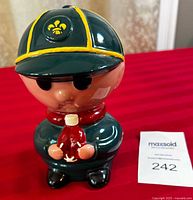 Front view of British Boy Scout ceramic piggy bank showing face, cap with emblem, and holding red bottle.