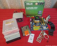 Photo showing the Asus M2N68-AM motherboard, three CD ROM drives stacked, Seagate 160GB hard drive, power supply unit, motherboard box and I/O shield on red surface