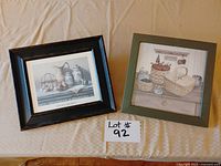 Two framed prints side by side; one with a basket of fruit in a green wood frame, the other with eggs and vegetables in a dark wood frame.