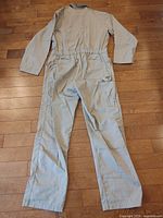 Back view of light gray federal worker coveralls laid flat on wooden floor showing long sleeves, elastic waist, and multiple pockets including back and side leg pocket.