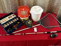 Full lot overview showing fishing rod with reel laid flat, open blue tackle box with lures and fishing tackle, white minnow bucket with net, orange trolling bait bucket, adjustable fishing net, and camo fishing hat arranged on red table.
