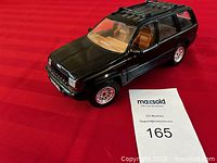 Black Jeep Grand Cherokee diecast model car featuring detailed tan interior, opening doors, and realistic chrome wheels on red background.