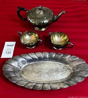 Full view of the silver teapot, cream and sugar containers, with the serving tray beneath, all placed on red cloth.