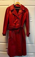 Front view of vintage red leather women's trench coat on a hanger against a white door, showcasing the coat's double-breasted button front, wide collar, and belt.
