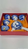 Full tea set of five cups and one teapot arranged in a red box on orange fabric, showing blue ceramic with cat motif.