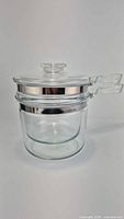 Full set of vintage Pyrex Flameware glass double boiler with two stacked pots and lid on top, showing clear glass and metal bands attached to rims and handles.
