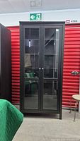 Front view of black IKEA Billy bookcase with glass paneled double doors, five shelves inside, and silver door knobs, standing in storage unit.
