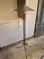 Full view of the modern matte black metal floor lamp standing on tiled floor next to white cabinetry and refrigerator. Shows the lamp's structure, legs, and cord.