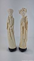 Photo showing both male and female Chinese-style faux ivory statuettes standing side by side on black bases.