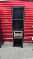 Full view of tall shelving unit showing three open shelves and two light wood-tone drawers with black handles in the center. Unit is black composite wood.
