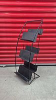 Side angled view of a black metal vintage bookstand showing its four slanted shelves and curved supporting frame against a red shutter background.
