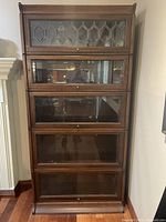 Front view of the antique wooden barrister bookcase with five glass-panelled shelves. Top section features decorative geometric leaded glass, below are four clear glass doors with knobs.