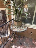 Full view of the mosaic patio table with lifelike silk plant placed on top, showing iron scroll legs and brown tiled floor background.