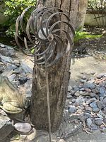Decorative garden windmill with spiral-shaped metal vanes mounted on a ground stake, set outdoors near a tree and stones.